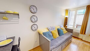 Uma sala de estar moderna com um sofá cinza, almofadas com padrão amarelo e azul, uma mesa de jantar com um prato amarelo e três relógios na parede.