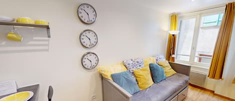 A modern living room with a grey sofa, yellow and blue patterned cushions, a dining table with a yellow plate, and three wall clocks.