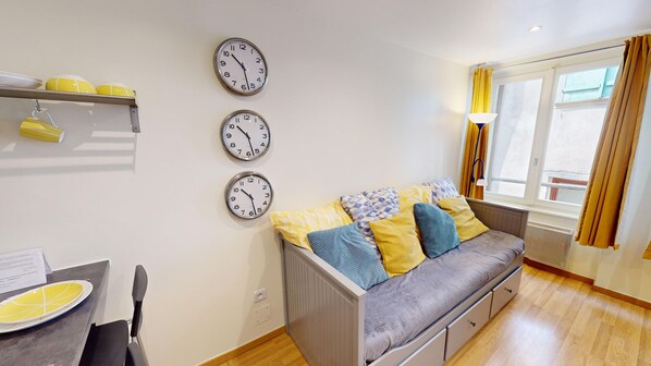 A modern living room with a grey sofa, yellow and blue patterned cushions, a dining table with a yellow plate, and three wall clocks.