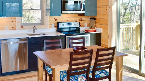 A wooden cabin kitchen with a dining area, wooden ceiling beams, and a large window.
