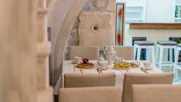 Mesa dispuesta con platos de comida, tazas y una tetera en una habitación con paredes de piedra y muebles blancos.