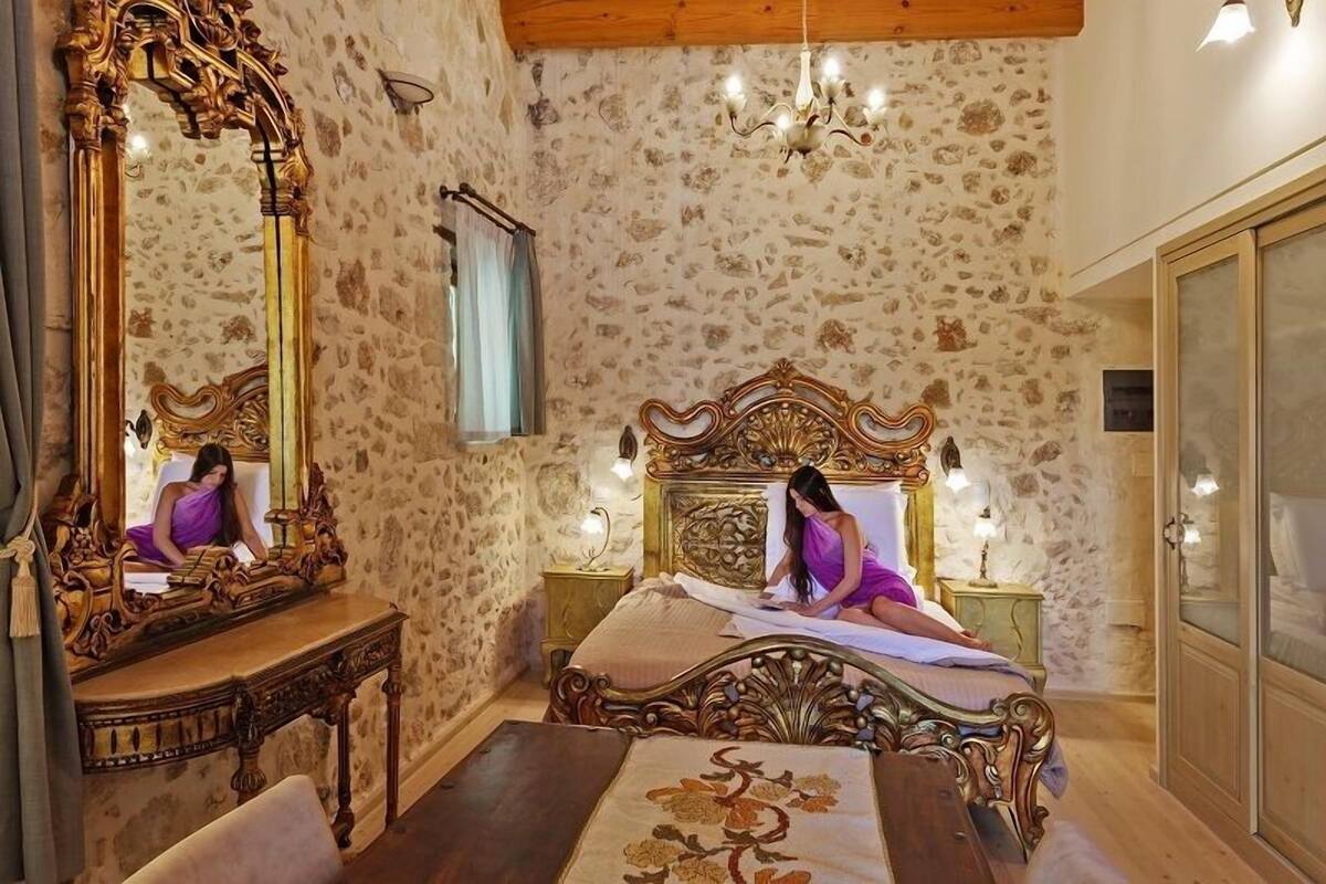 Un dormitorio con pared de piedra, una cama grande y elaborada, una mesita de noche de madera, un espejo y una araña.