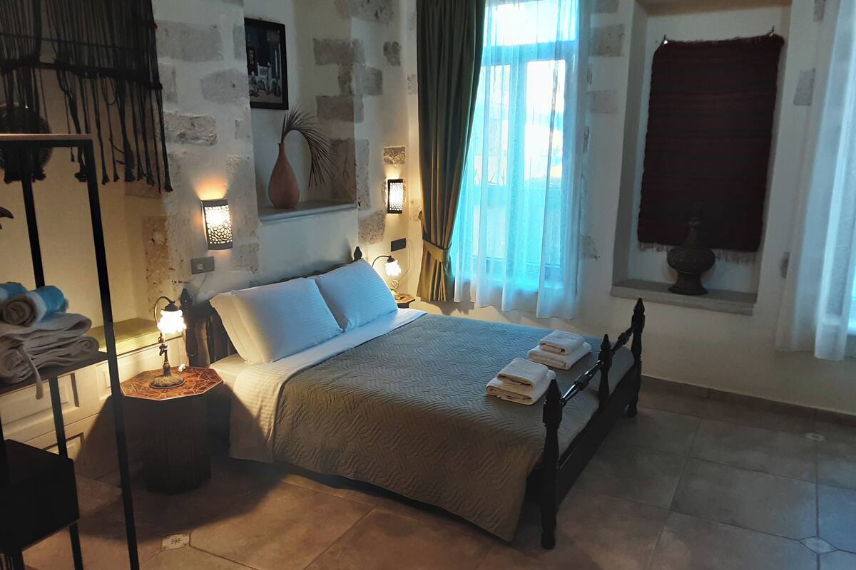 Un dormitorio con cama, mesitas de noche, lámpara, un florero y una ventana con cortinas.