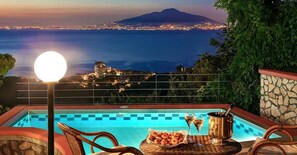 Une vue sur la piscine, avec une vue dégagée sur la mer et une montagne en arrière-plan, une chaise en osier, une table avec des collations et des boissons, et un lampadaire.