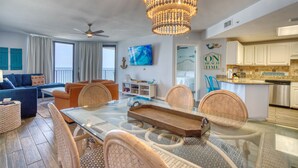 A spacious living room with a glass dining table, wicker chairs, a chandelier, and a view of the ocean.