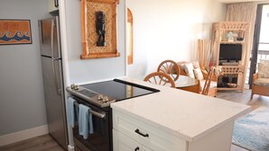 A kitchen with a stove, oven, and white cabinets. There is a dining area with wicker chairs and a table. - Beautiful Partial Ocean View Condo with Tropical Furnishings (Kihei)