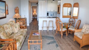 A living room with a sofa, a coffee table, and wicker chairs. There is a kitchen area with a counter and stools. - Beautiful Partial Ocean View Condo with Tropical Furnishings (Kihei)