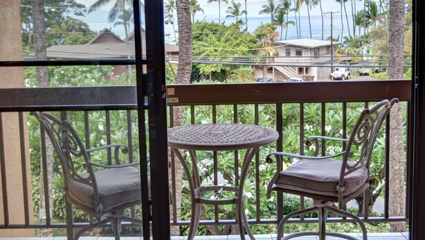 A balcony with two chairs and a small table, overlooking a residential area with palm trees and a view of the ocean. - Beautiful Partial Ocean View Condo with Tropical Furnishings (Kihei)