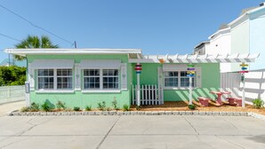 Una piccola casa a un piano con facciata verde e dettagli bianchi. Dispone di un portico con recinzione in legno bianco e un piccolo giardino con piante e una panchina.