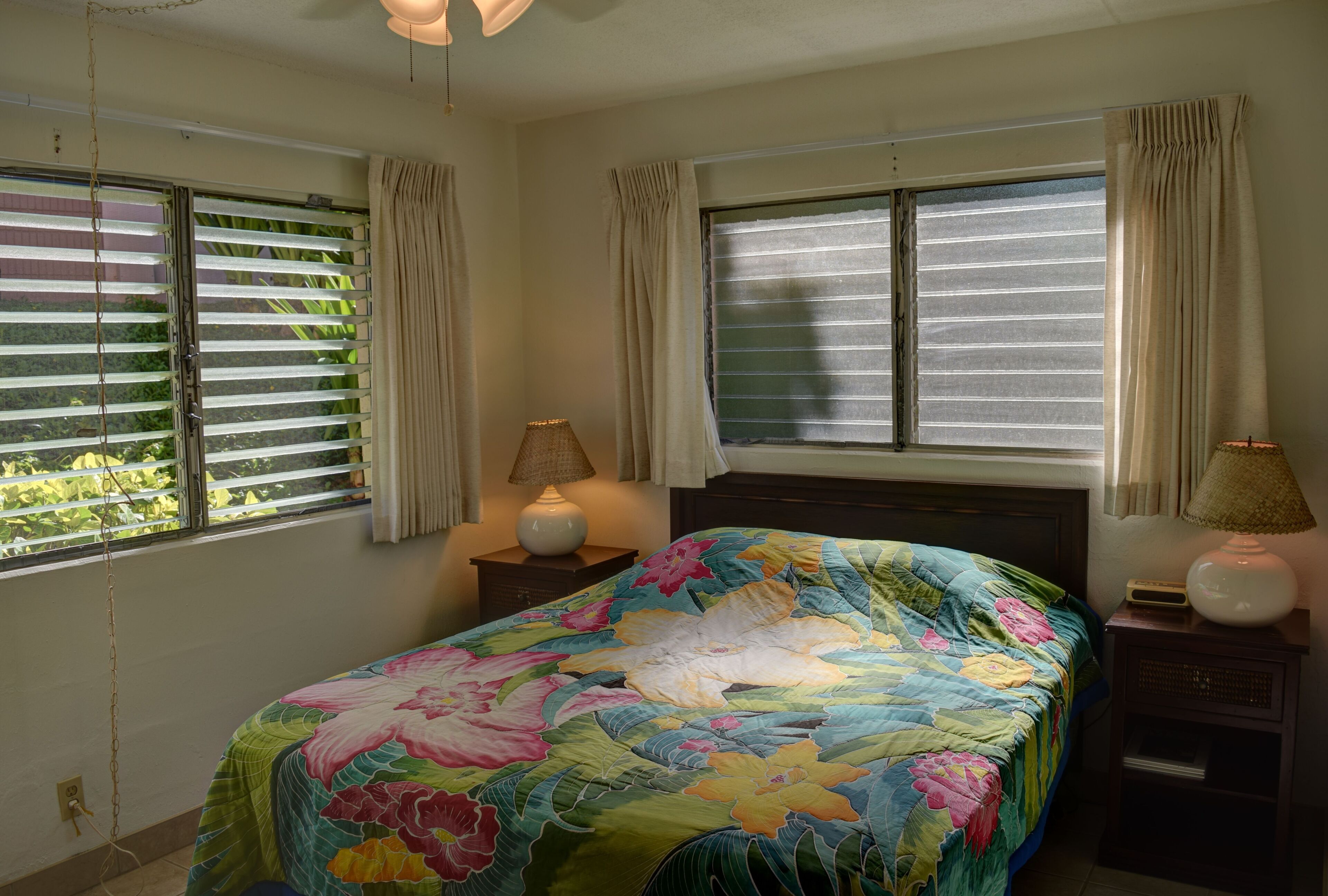 Une chambre à coucher avec une couvre-lit à motifs floraux, deux lampes de chevet et des rideaux aux deux fenêtres.