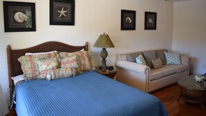 A bedroom with a bed, a nightstand, a lamp, a sofa, and framed pictures on the wall. - Bright and Spacious Corner Studio Unit with Wrap Around Lanai (Kihei)