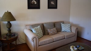 A living room with a beige sofa, two framed pictures on the wall, a lamp on a side table, and a remote control on the floor. - Bright and Spacious Corner Studio Unit with Wrap Around Lanai (Kihei)