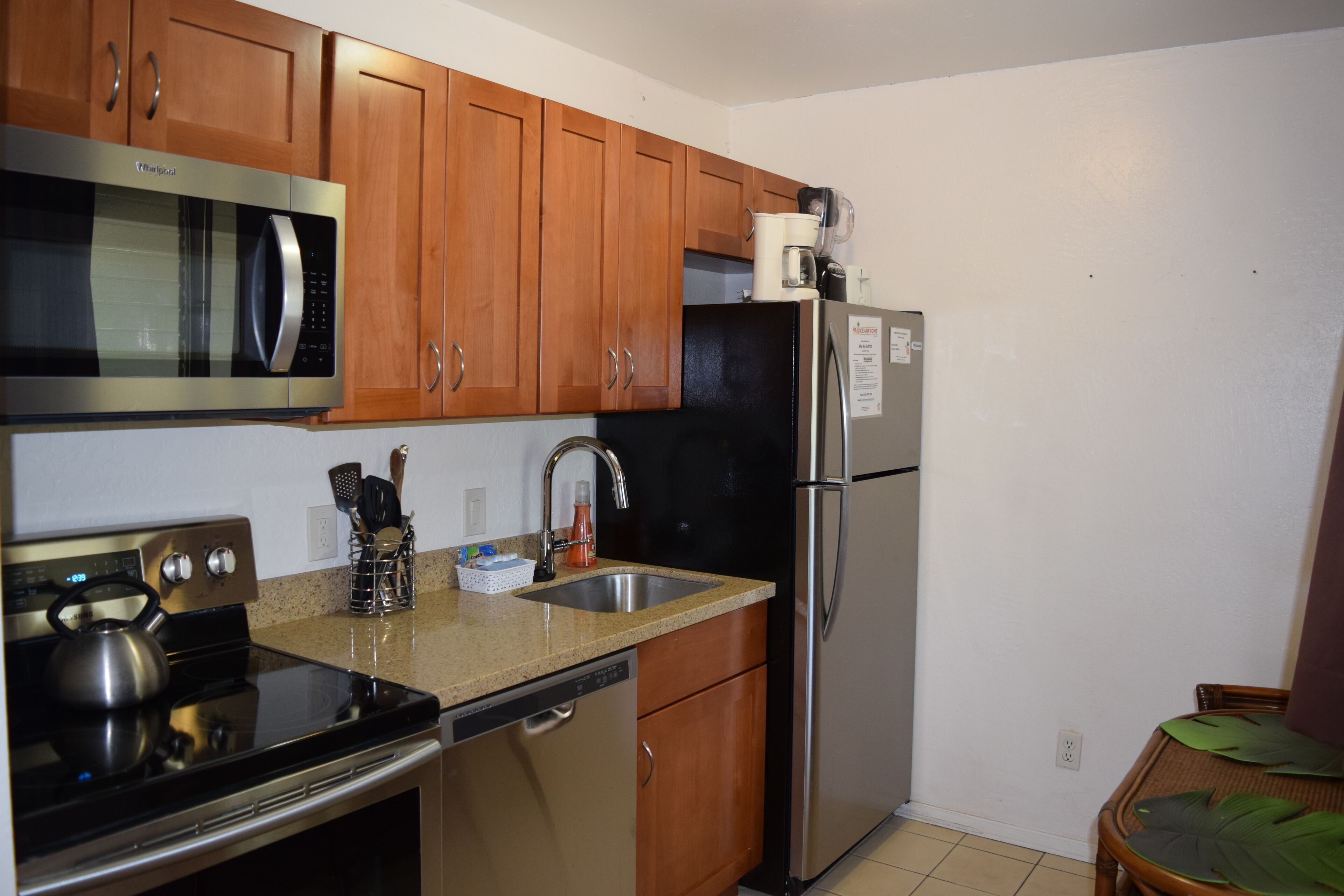 A kitchen with wooden cabinets, a stainless steel microwave, a refrigerator, a sink, and a kettle on the stove.