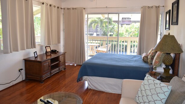 A bedroom with a large bed, wooden dresser, and a balcony with a view of palm trees. - Bright and Spacious Corner Studio Unit with Wrap Around Lanai (Kihei)