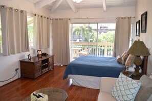 A bedroom with a bed, a dresser, a chair, a table, and a balcony with a view of palm trees.