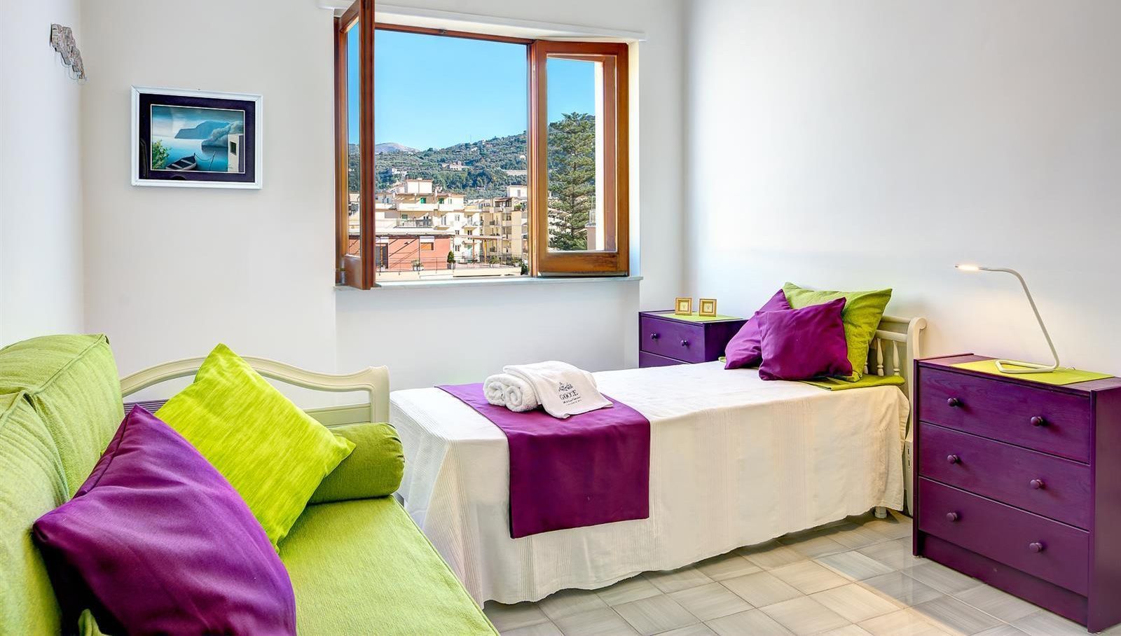Quarto com cama, penteadeira, mesa de cabeceira, janela com vista para edifícios e quadro na parede.