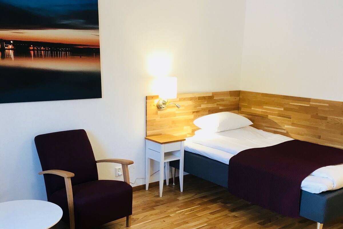 A hotel room with a wooden floor, a bed with a wooden headboard, a nightstand with a lamp, a chair, a small table, and a colorful rug.
