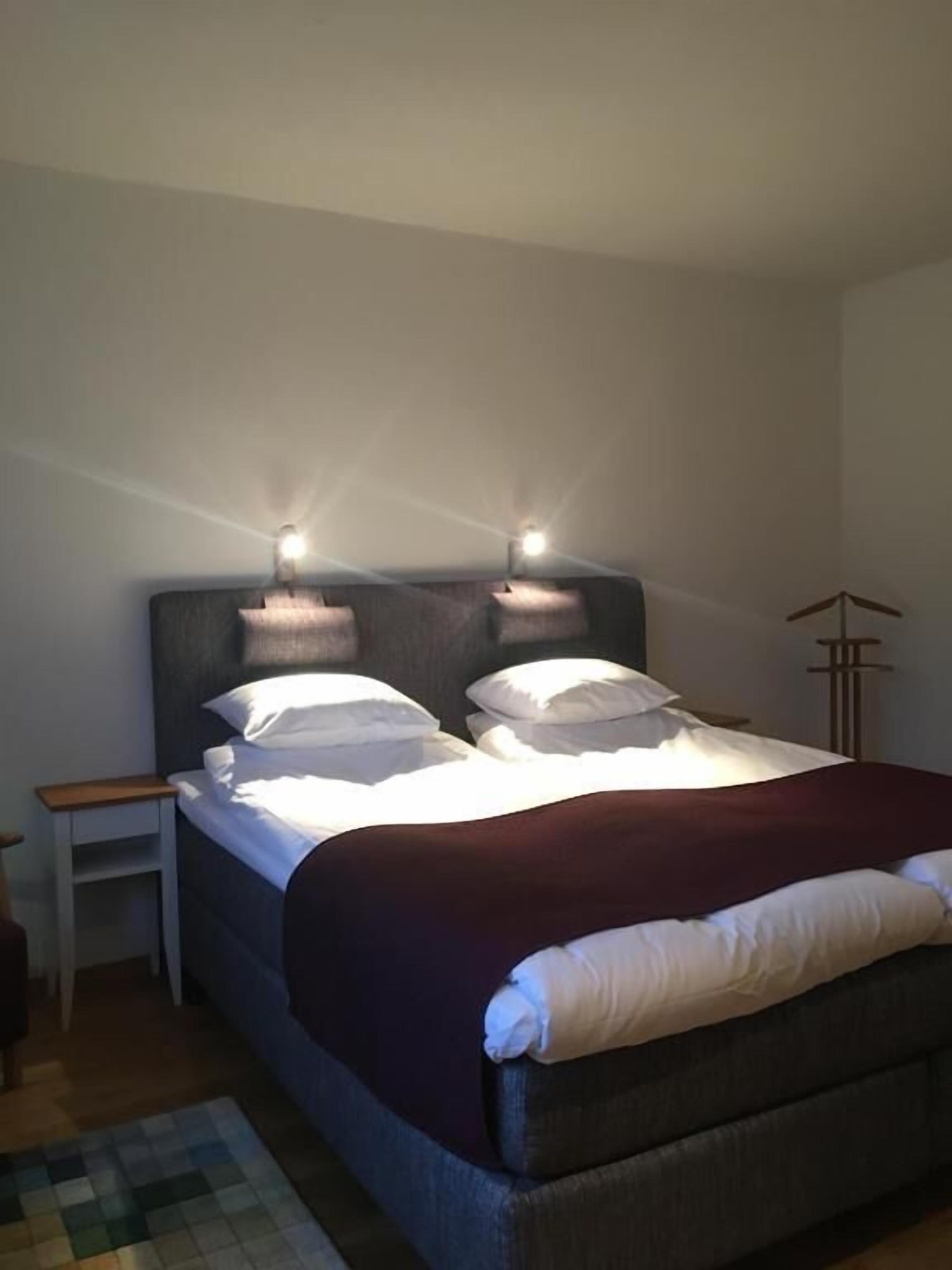 Una cama bien hecha con sábanas blancas y un cabecero oscuro, junto con una mesita de noche y una lámpara de pie.
