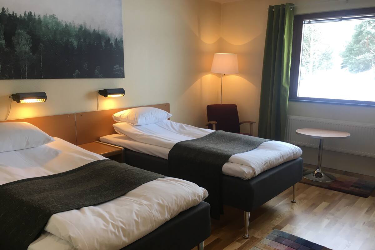 A hotel room with two beds, a nightstand, a chair, a table, and a painting on the wall.