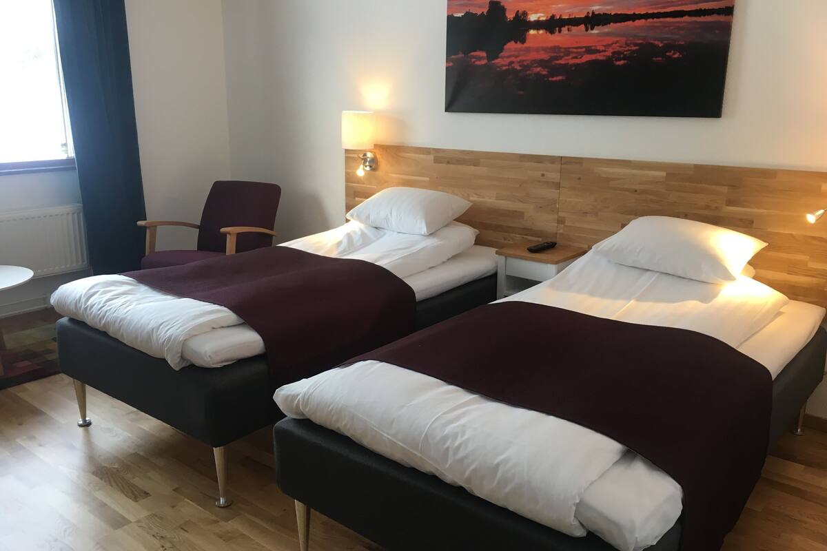A hotel room with two beds, a chair, a table, and a painting of a sunset.