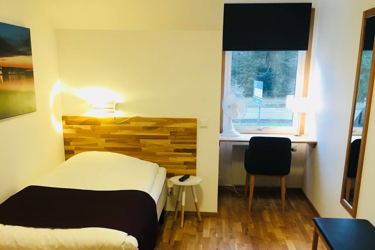 A hotel room with a wooden headboard bed, a desk with a chair, a small stool, and a colorful rug.