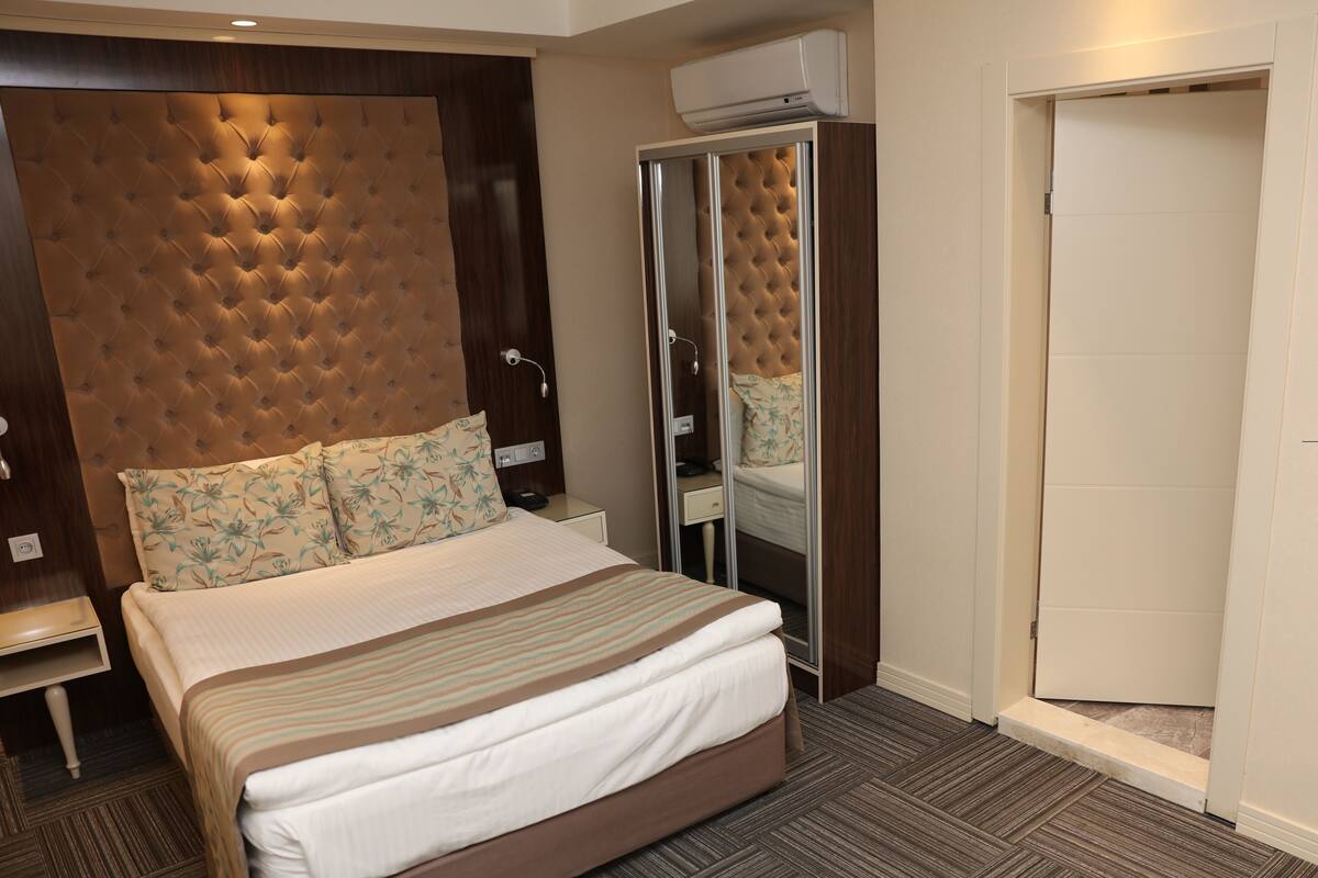 Habitación de hotel con una cama grande, un armario con espejo, una mesita de noche y un equipo de aire acondicionado.