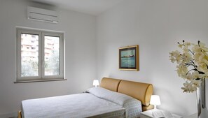 Ein Schlafzimmer mit einem Bett, zwei Nachttischlampen, einem gerahmten Bild an der Wand und einem Fenster mit Blick auf Gebäude.