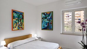 Ein Schlafzimmer mit einem Bett, zwei Bildern, einem Fenster und einem Nachttisch mit einer Lampe.