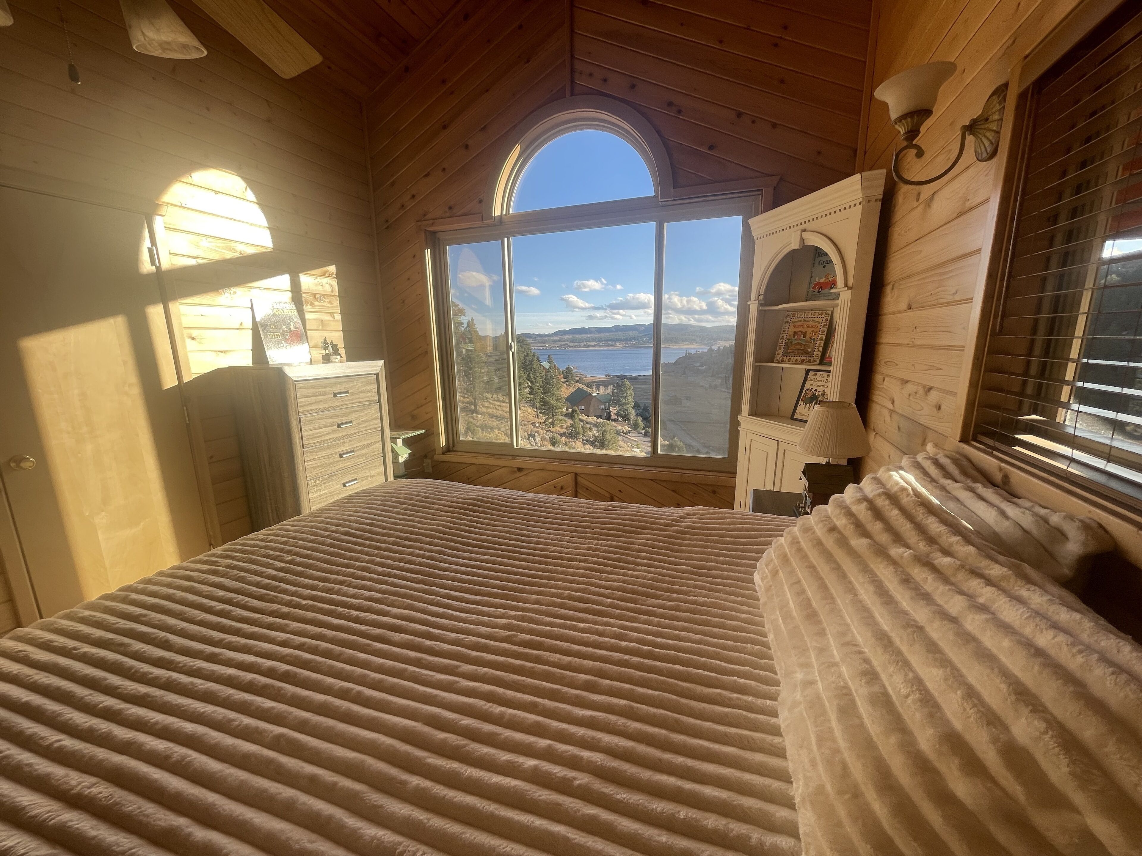 A bedroom with a large bed, a dresser, a vanity, and a window with a view of a lake and mountains.