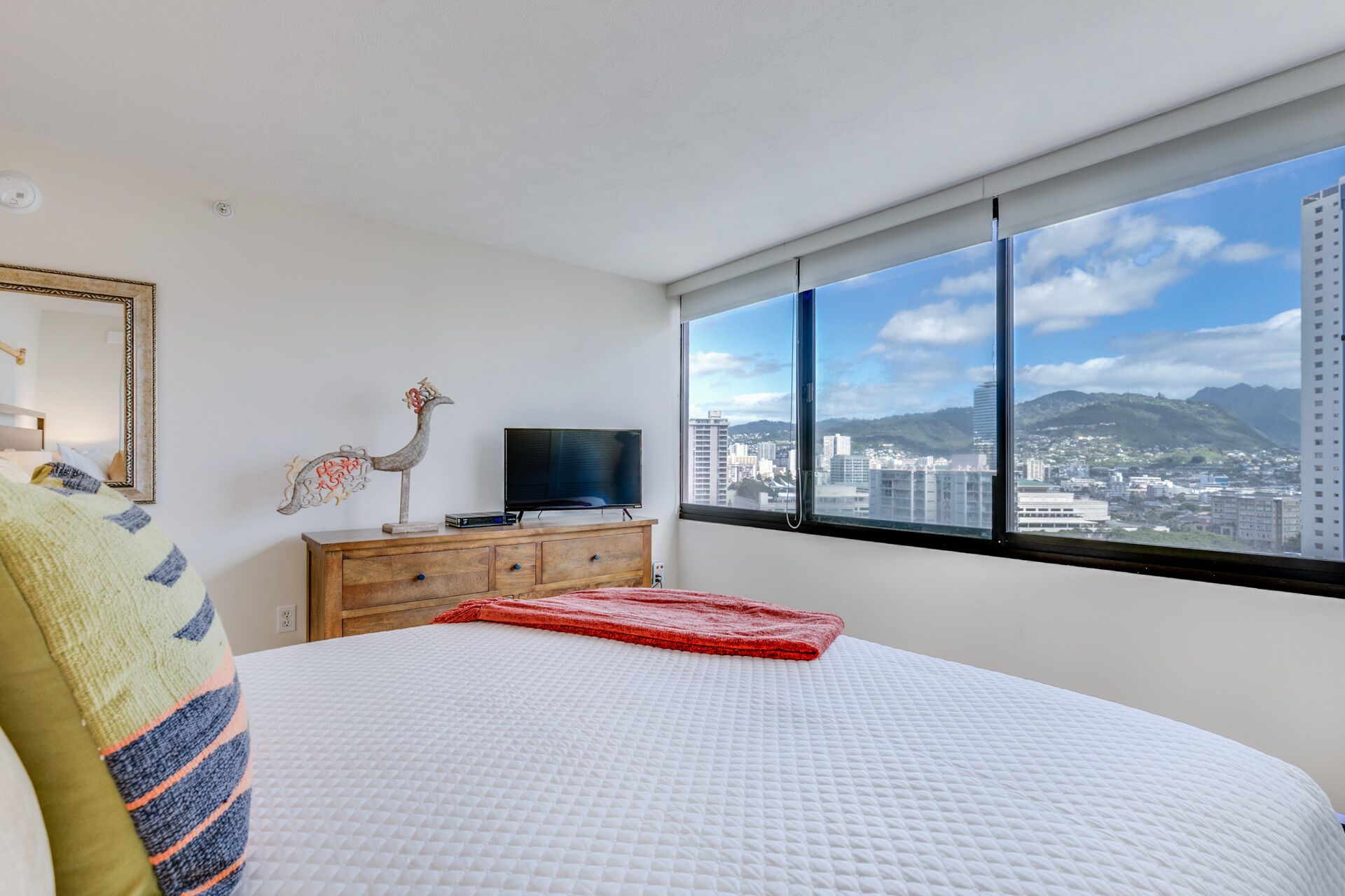 Ein Schlafzimmer mit einem großen Bett, einer Holzkommode, einem Fernseher und Blick auf die Stadt durch ein großes Fenster.