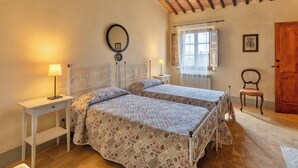 Ein Schlafzimmer mit zwei Betten, einem Nachttisch mit Lampe, einem Sessel, einer Holztür und einem Fenster mit Vorhängen.
