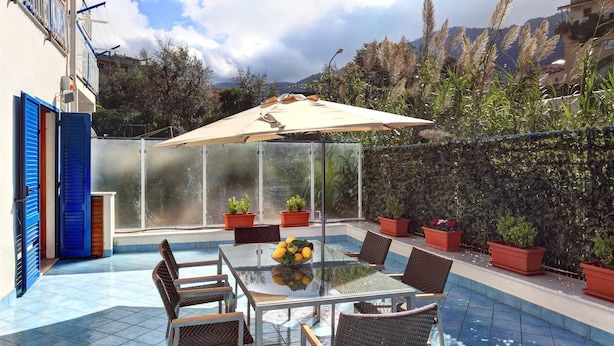 Une terrasse fermée par des baies vitrées, avec une table et des chaises, entourée de plantes en pot et d’une piscine.