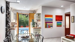 Une cuisine moderne avec des meubles blancs, un coin repas doté d’une table en verre et une vue sur un jardin par une porte ouverte.