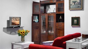 Un salon avec un canapé rouge, une table basse blanche, un téléviseur posé sur un meuble, un buffet en bois avec des portes en verre et un vase rempli de tournesols.