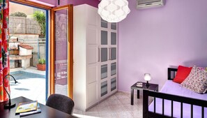 Une chambre comprenant un lit, un bureau avec une lampe, une chaise, une petite table avec une lampe, et une porte-fenêtre coulissante donnant sur un espace extérieur avec une terrasse et une vue sur un jardin.