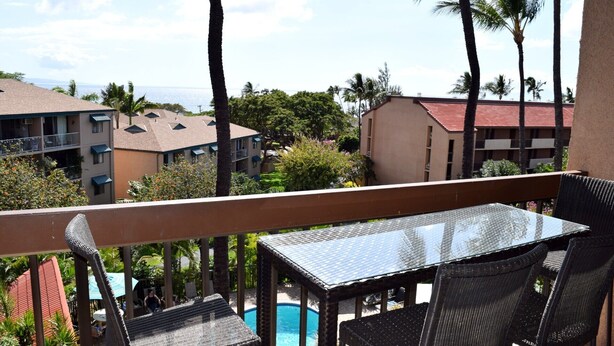 A balcony with a table and chairs overlooking a swimming pool, surrounded by palm trees and buildings. - Beautiful, Updated Condo Across the Street from Famous Charley Young Beach (Kihei)