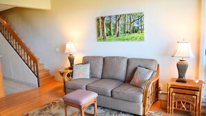 A living room with a grey sofa, two wooden side tables, a wicker stool, a staircase, and a framed forest painting on the wall. - Beautiful, Updated Condo Across the Street from Famous Charley Young Beach (Kihei)