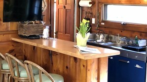 A wooden interior with a bar area, wooden chairs, a television, and a kitchen. - Compass Rose Cottage ★ Steps to Flax Pond & Nature Trails ★ Kayaks  (Dennis)