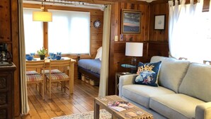 A cozy room with a wooden floor, a sofa, a small table, a dining area with chairs, and a bed. - Compass Rose Cottage ★ Steps to Flax Pond & Nature Trails ★ Kayaks  (Dennis)