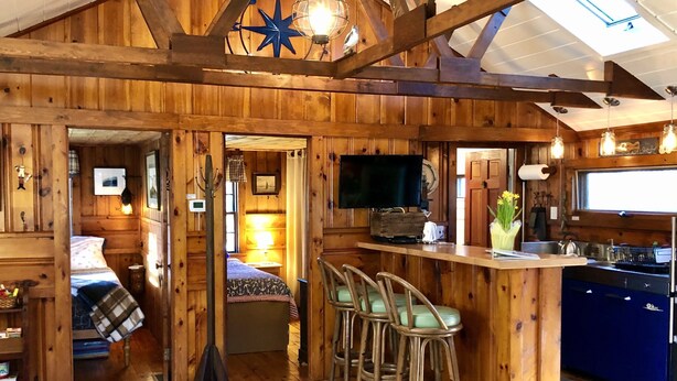A rustic cabin interior with a wooden bar area, a television, and a kitchen. - Compass Rose Cottage ★ Steps to Flax Pond & Nature Trails ★ Kayaks  (Dennis)