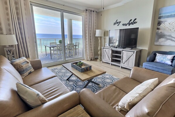 A modern living room with a large sofa, a coffee table, and a TV. There is a balcony with a view of the sea.