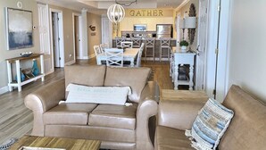 Amplia sala de estar con un sofá color beige, una mesa de centro y un comedor con mesa y sillas blancas. Se observa una cocina con gabinetes blancos y una lámpara de candelabro al fondo.
