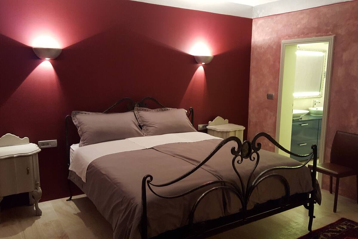 A bedroom with a bed, two bedside tables, a chair, and a bathroom visible through an open door.