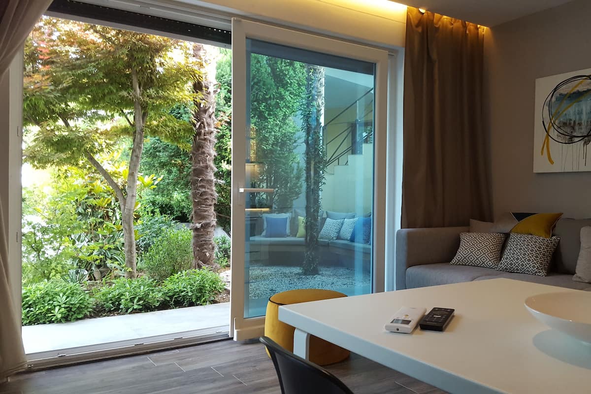 A modern living room with a glass door leading to a garden, a sofa with patterned cushions, and a minimalist table with a bowl.