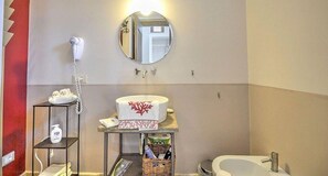 Kylpyhuoneessa on valkoinen pesuallas, pyöreä peili, hiustenkuivaaja, hylly, jossa on hygieniatuotteita, ja kori, jossa on pyyhkeitä.