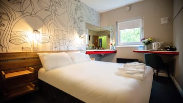 Ein Hotelzimmer mit einem großen Bett, einem hölzernen Kopfteil, einem Schreibtisch mit Stuhl, einem Fenster und einer Wand mit Spiegel und Wandbild.