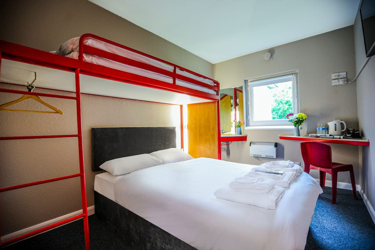 A hotel room with a bunk bed, a desk with a chair, a window with a view, and a bed with neatly made sheets.