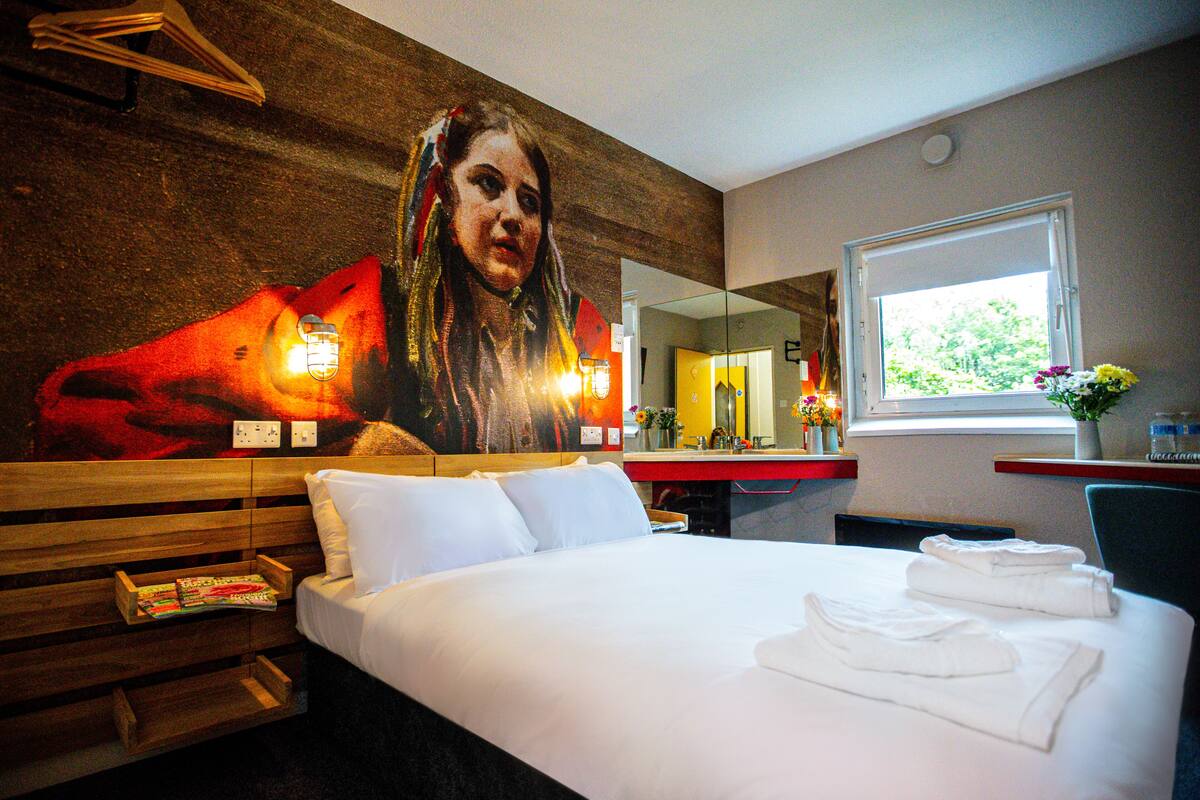 A hotel room with a large bed, a wooden headboard, a mirror, a window, and a wall with a mural.