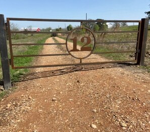 Una reja de metal con el número 12, un sendero de tierra y un paisaje rural con un granero rojo al fondo.