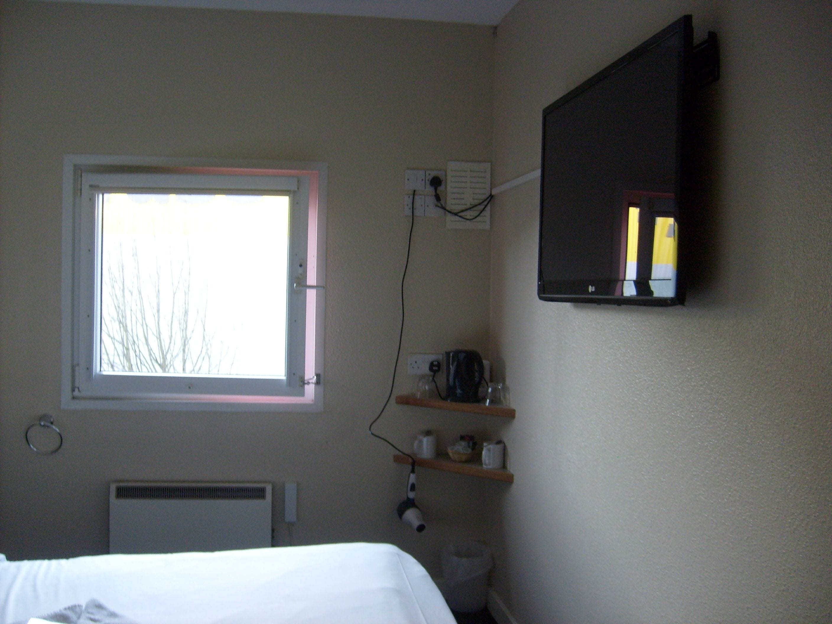 Un dormitorio con ventana, televisor de pared, radiador y estante con artículos de aseo.